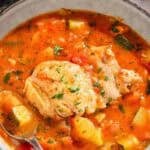 A blow of the chicken stew with a spoon next to a cloth napkin, pieces of crusty bread, a bowl of chopped parsley, and the rest of the stew in a large pot.