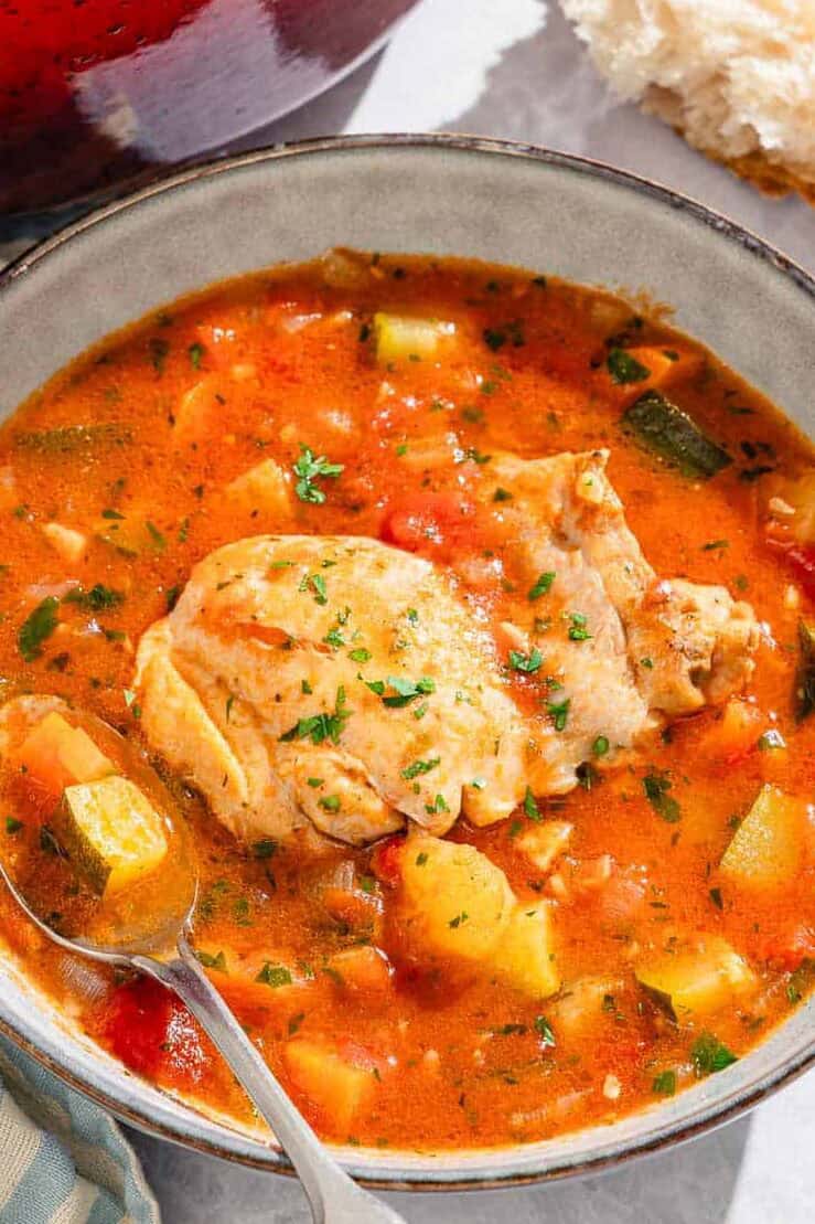 A blow of the chicken stew with a spoon next to a cloth napkin, pieces of crusty bread, a bowl of chopped parsley, and the rest of the stew in a large pot.