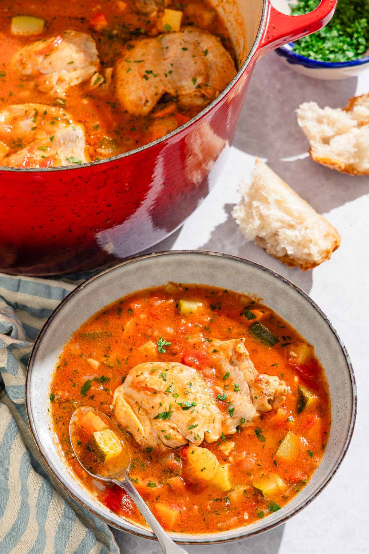 A blow of the chicken stew with a spoon next to a cloth napkin, pieces of crusty bread, a bowl of chopped parsley, and the rest of the stew in a large pot.