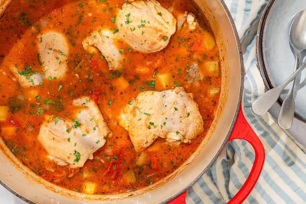 Chicken stew in a large pot next to a stack of 2 bowls with 2 spoons.