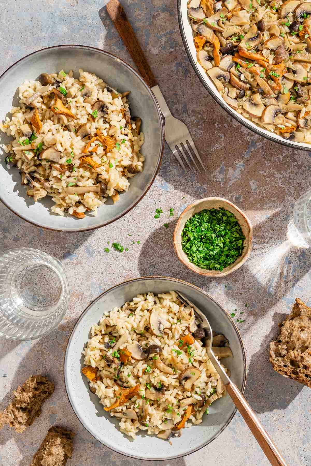 Two servings of mushroom rice in a bowl, one with a spoon. Next to these are pieces of bread, another fork, a glass of water, a bowl of chopped parsley, and the rest of the mushroom rice in a skillet.