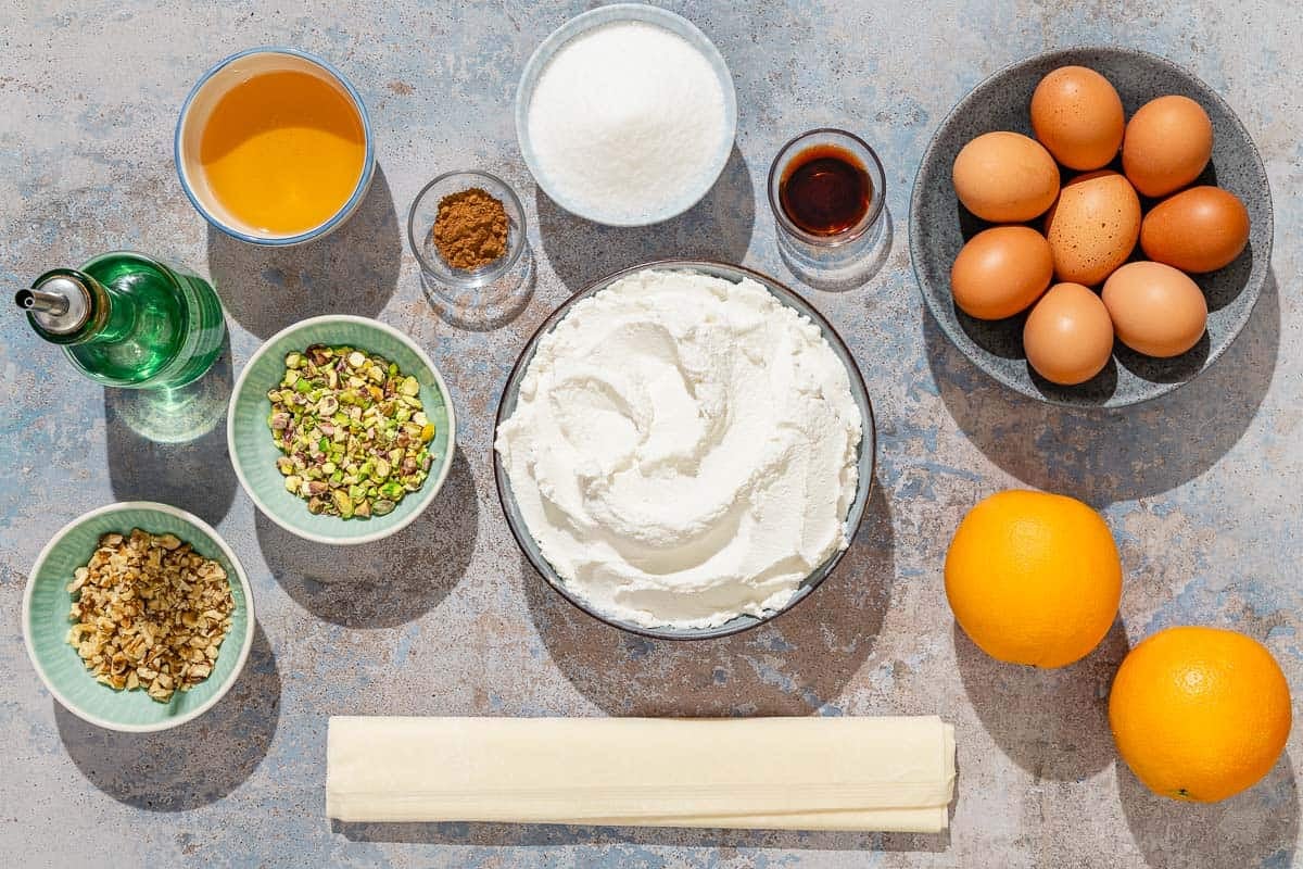 Ingredients for baklava cheesecake including ricotta cheese, eggs, sugar, vanilla extract, oranges, olive oil, walnuts, pistachios, cinnamon, phyllo dough, and honey.