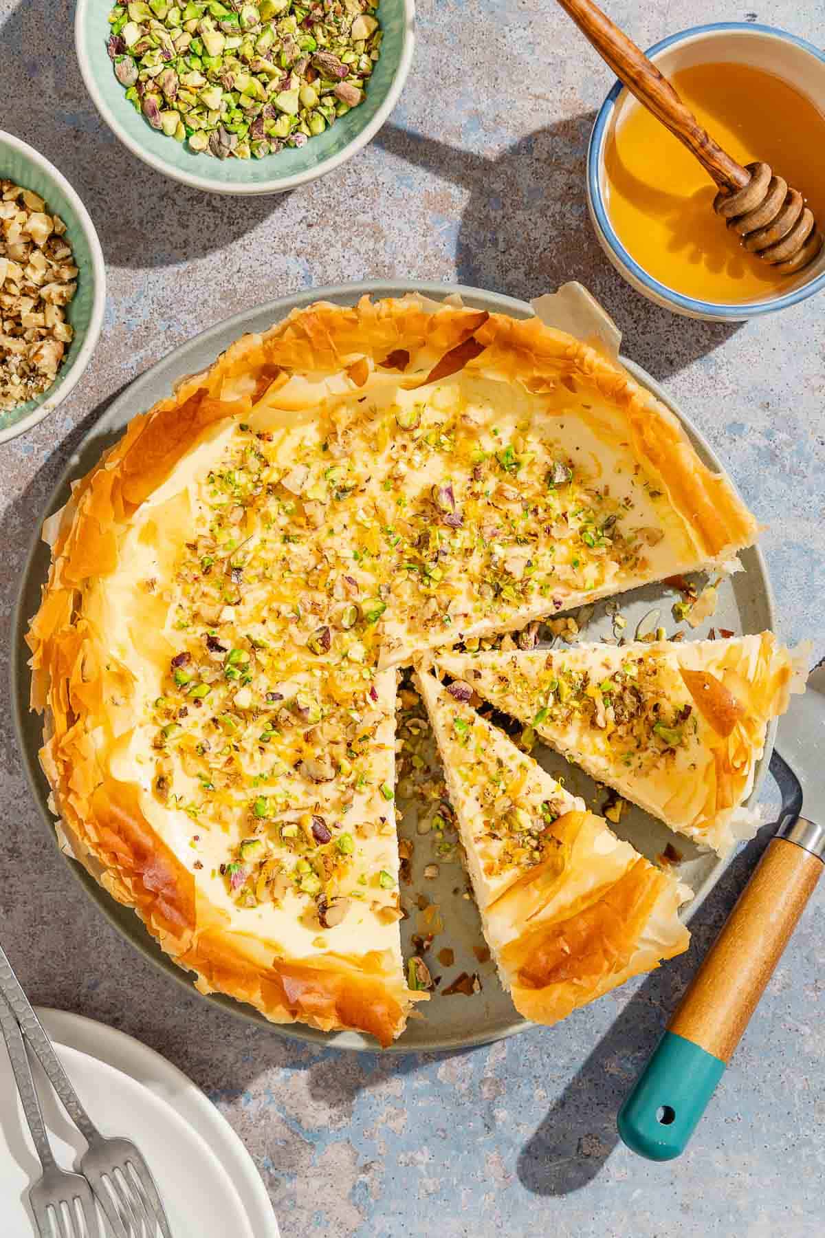 An overhead photo of 2 slices of baklava cheesecake on a platter with the rest of the cake. Surrounding this are bowls of nuts, a stack of 2 plates with 2 forks, a bowl of honey with a honey dipper, and a cake serving utensil.