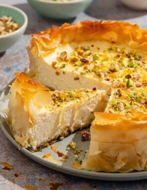 A close up of a slice of baklava cheesecake on a platter with the rest of the cake. I the background are small bowls of nuts.