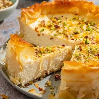 A close up of a slice of baklava cheesecake on a platter with the rest of the cake. I the background are small bowls of nuts.
