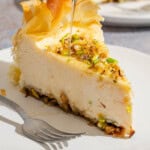 Honey being drizzled on a slice of baklava cheesecake on a plate with a fork.