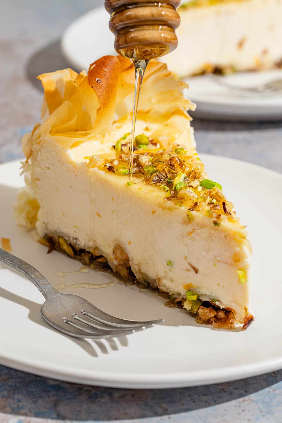 Honey being drizzled on a slice of baklava cheesecake on a plate with a fork.