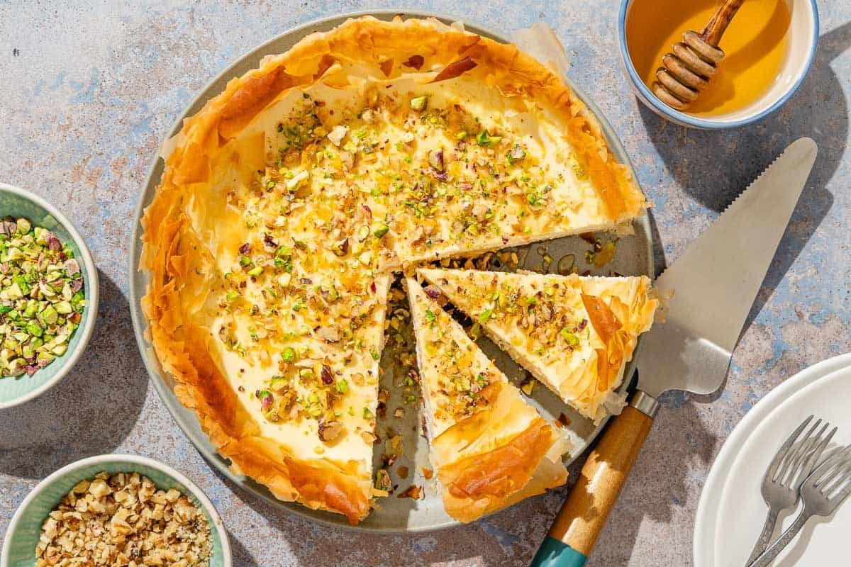 An overhead photo of 2 slices of baklava cheesecake on a platter with the rest of the cake. Surrounding this are bowls of nuts, a stack of 2 plates with 2 forks, a bowl of honey with a honey dipper, and a cake serving utensil.