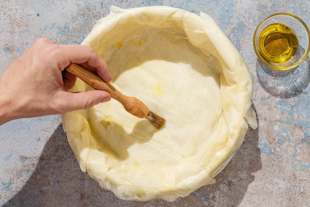 Olive oil being brushed on the phyllo crust for the baklava cheesecake. Next to this is a bowl of olive oil.