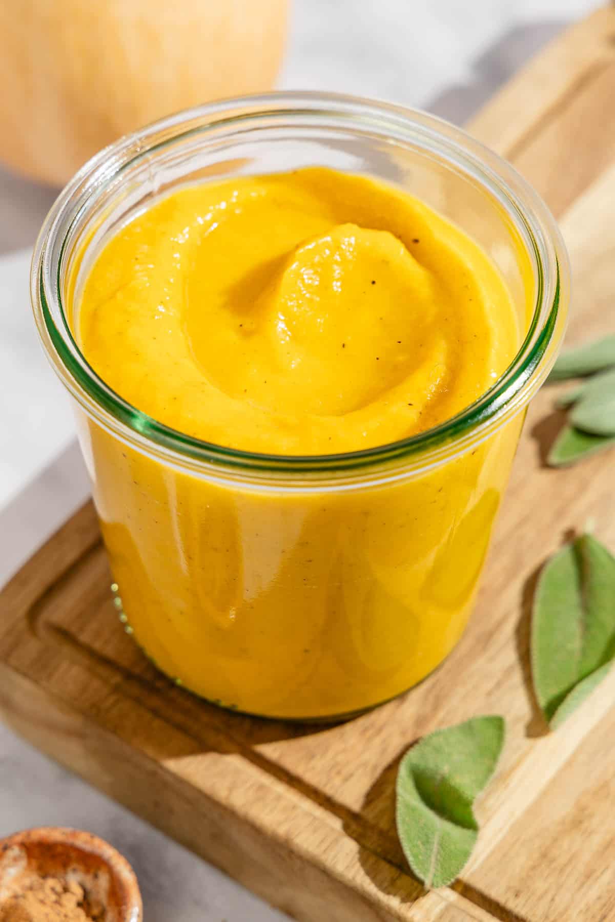 Butternut squash pasta sauce in a jar on a cutting board with a few sage leaves. Next to this is a bowl of ground nutmeg and a butternut squash.
