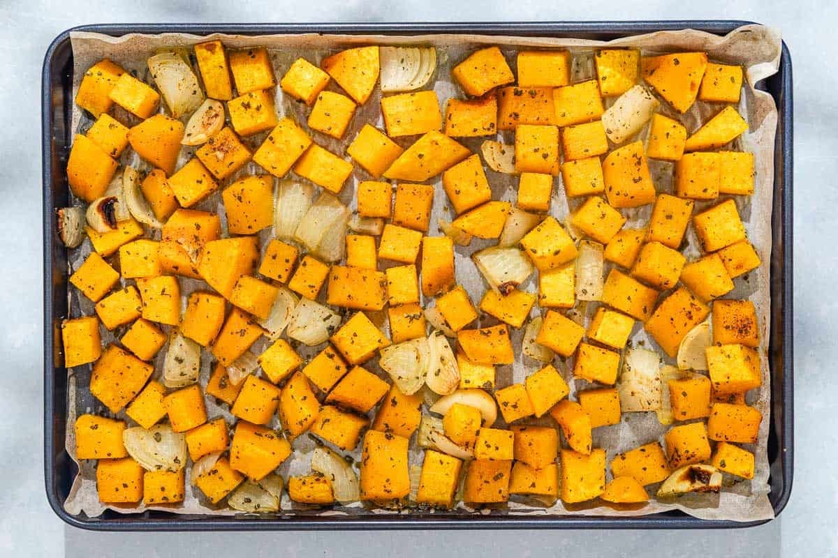 Roasted pieces of butternut squash, onion and garlic on a parchment lined sheet pan.