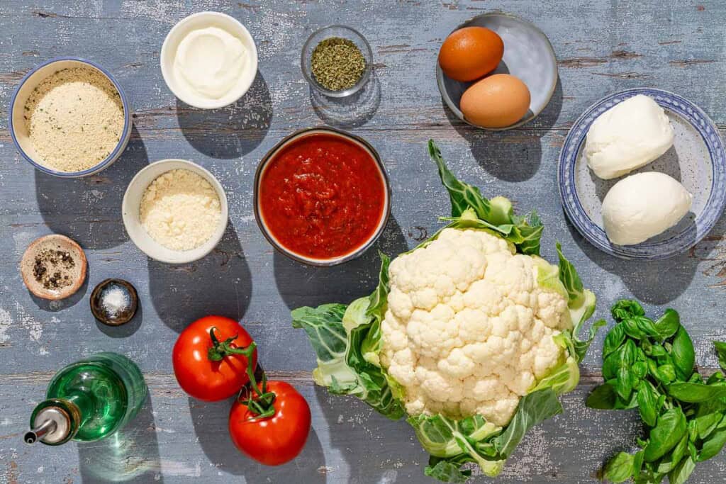 Ingredients for cauliflower parmesan including cauliflower, salt, black pepper, eggs, greek yogurt, dried oregano, breadcrumbs, grated parmesan cheese, olive oil, marinara sauce, tomatoes, and mozzarella cheese.