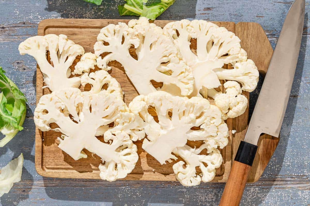 Five cauliflower slices on a cutting board with a knife.