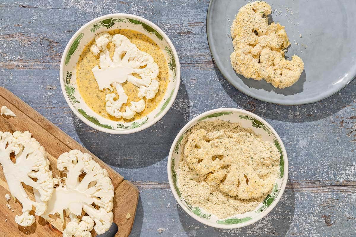 A cutting board with 2 slices of cauliflower next to a bowl of the batter with one cauliflower slice, a bowl of the breadcrumbs with a cauliflower slice, and a breaded cauliflower slice on a platter.