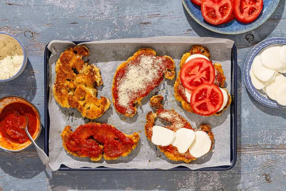 The battered and fried cauliflower slices on a parchment lined sheet pan. Each is partially topped with the rest of the baked cauliflower parmesan ingredients. Surround this sheet pan is a bowl of marinara with a spoon, bowl of the bread crumbs, a plate of sliced tomatoes, and a plate of sliced mozzarella cheese.