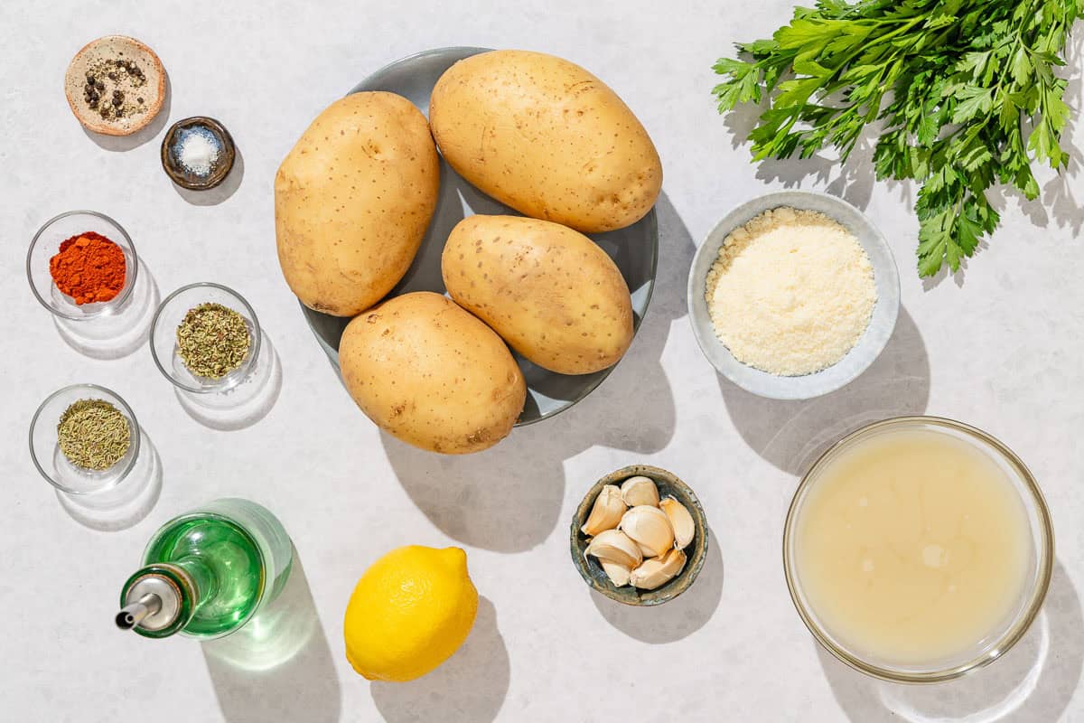 Ingredients for greek potatoes including baking potatoes, salt, pepper, paprika, rosemary, oregano, garlic, olive oil, lemon, broth, grated parmesan, and parsley.