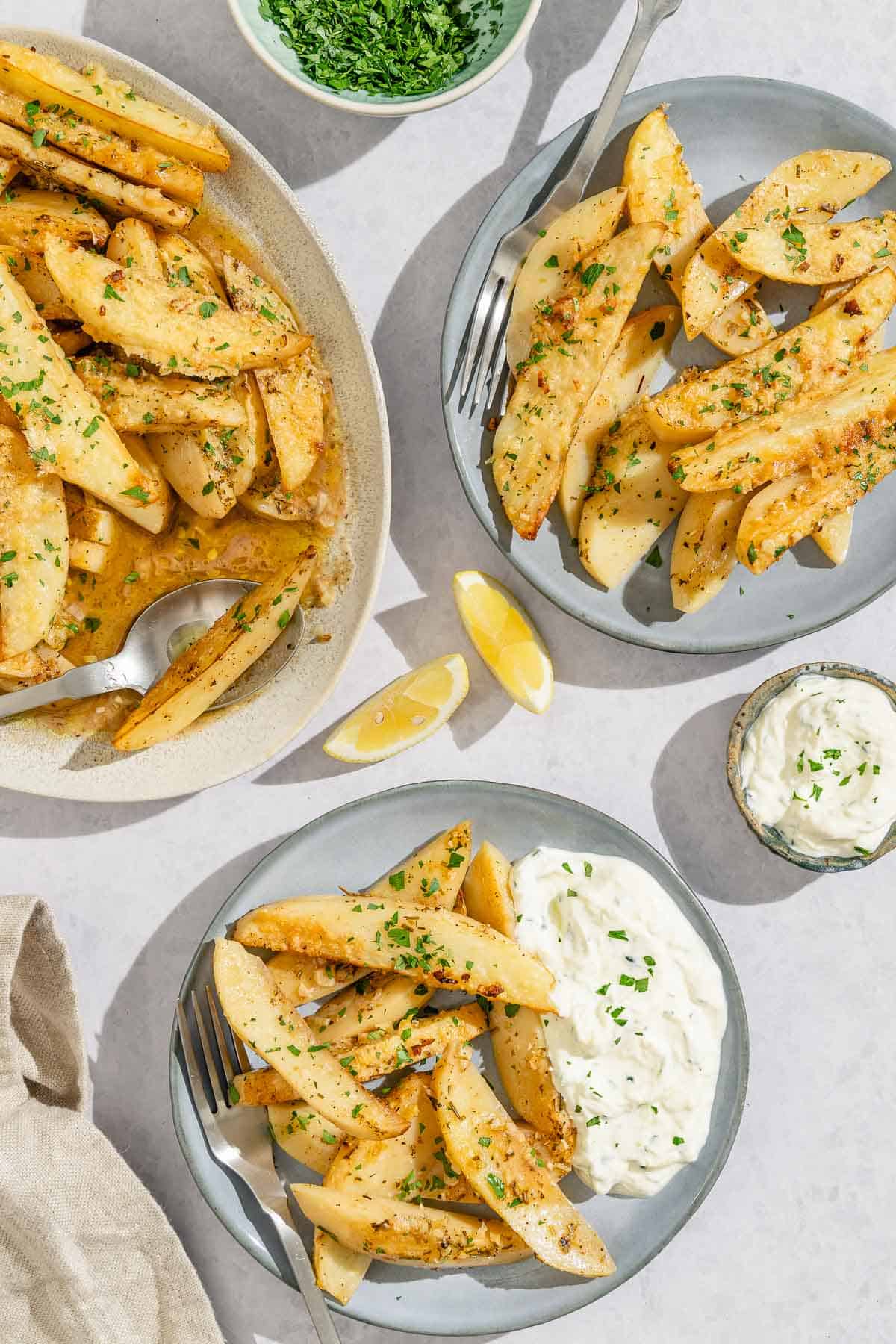 Two plates of the greek lemon potatoes on plates with forks, one with a side of tzatziki. Next to these are bowls of chopped parsley and tzatziki, 2 lemon wedges and the rest of the greek potatoes on a platter with a serving spoon.