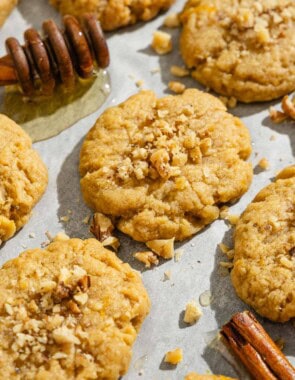 A close up of several melomakarona on a parchment lined sheet pan with a cinnamon stick and a honey dipper with a bit of honey.
