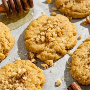 A close up of several melomakarona on a parchment lined sheet pan with a cinnamon stick and a honey dipper with a bit of honey.
