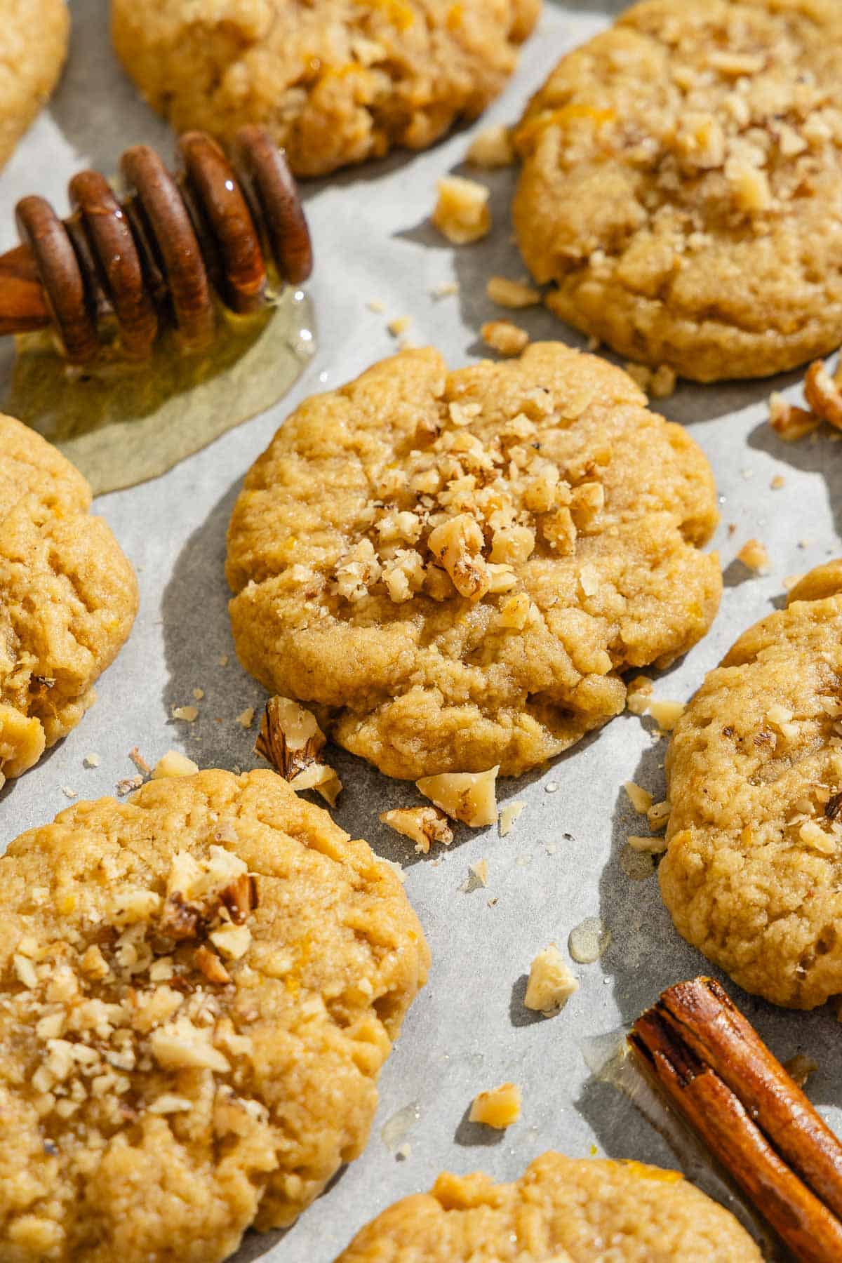 A close up of several melomakarona on a parchment lined sheet pan with a cinnamon stick and a honey dipper with a bit of honey.