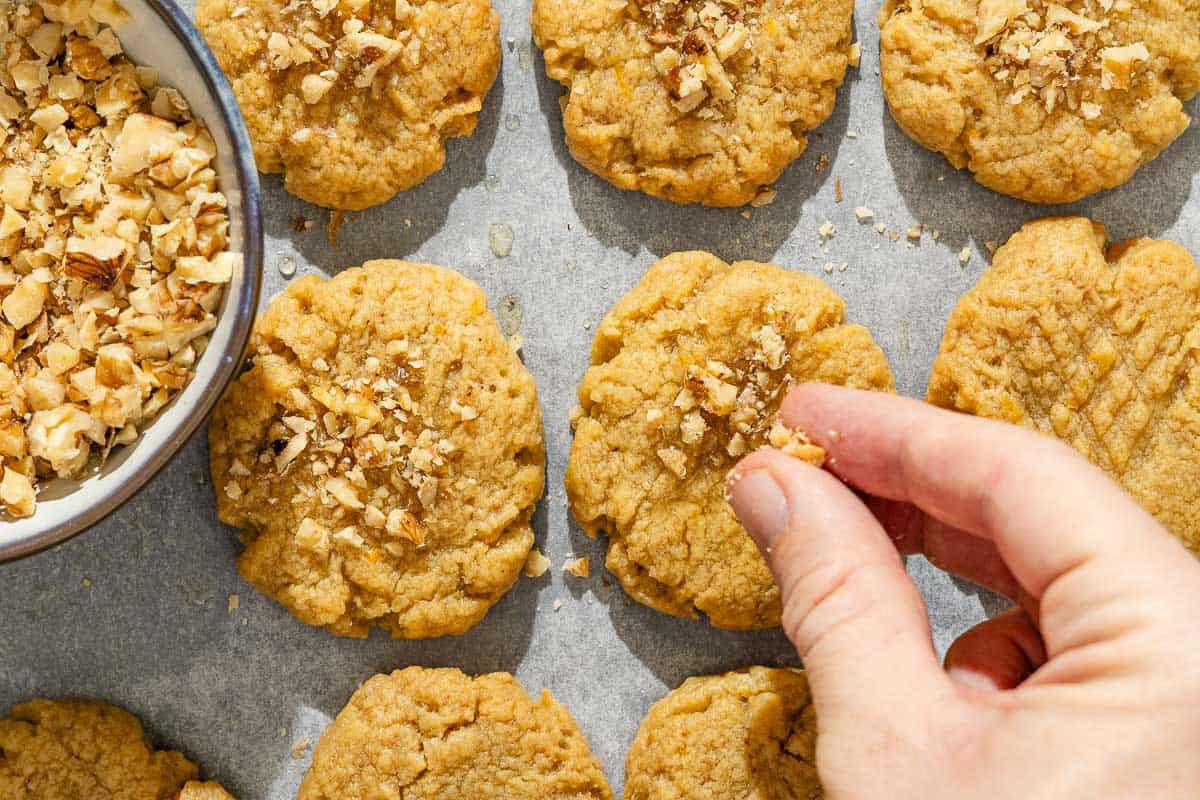 The chopped walnuts being added to the melomakarona that are spread evenly on a parchment lined sheet pan. Next to the cookies is a bowl of the chopped walnuts.