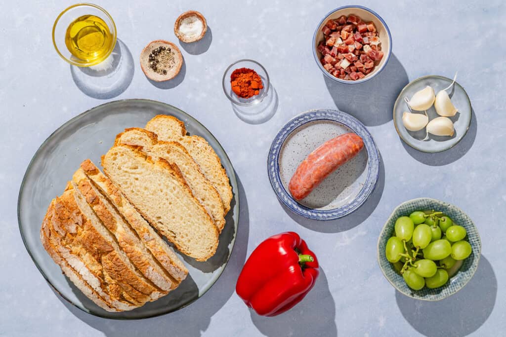 Ingredients for migas including crusty bread, olive oil, chorizo, pancetta, garlic, red bell pepper, sweet paprika, grapes, salt and pepper.