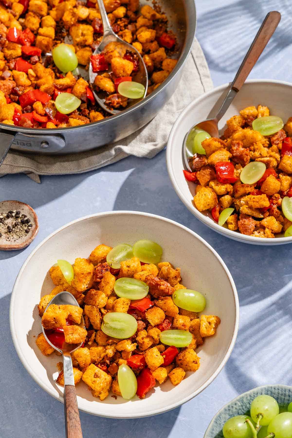 Two bowls of migas with spoons next to the rest of the migas in a tall sided skillet with a serving spoon, a bowl of pepper and a bowl of grapes.