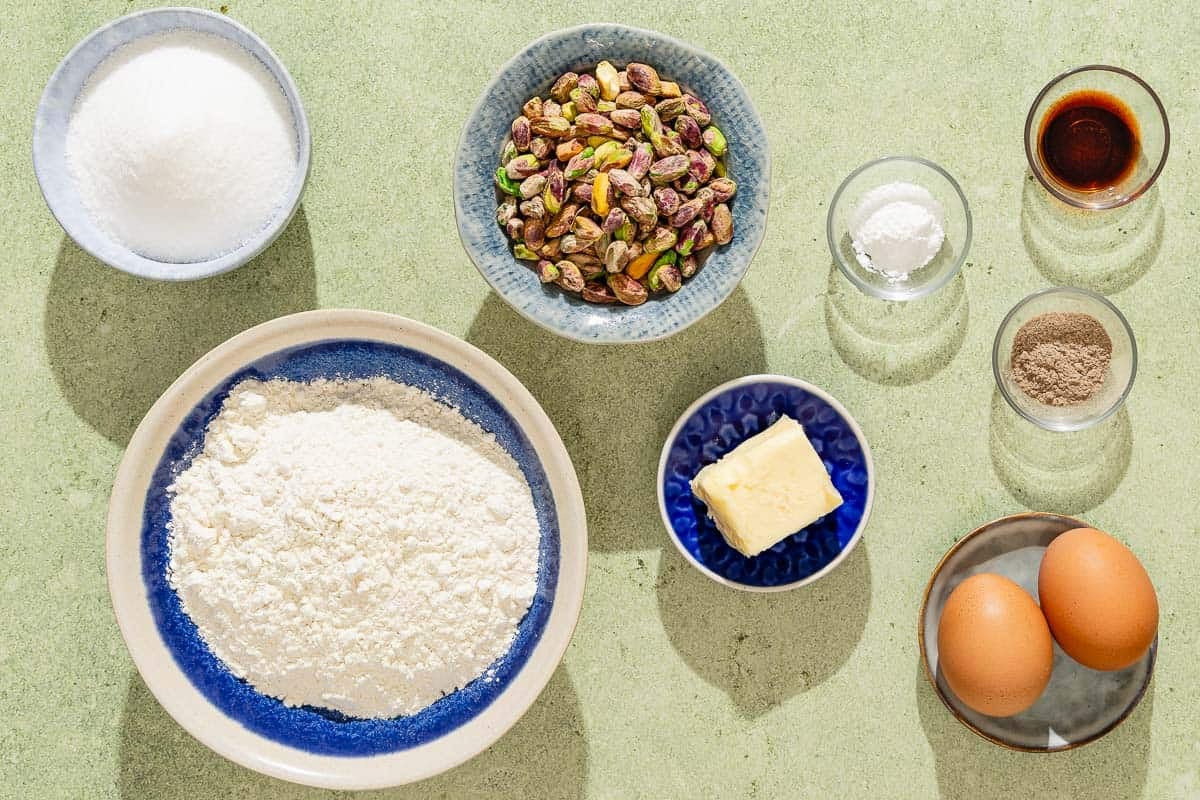 Ingredients for pistachio biscotti including flour, baking powder, butter, sugar, eggs, pistachios, ground green cardamom and vanilla extract.