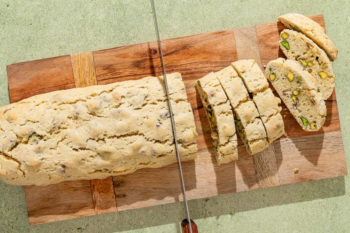 The baked pistachio biscotti being sliced on a cutting board with a knife.