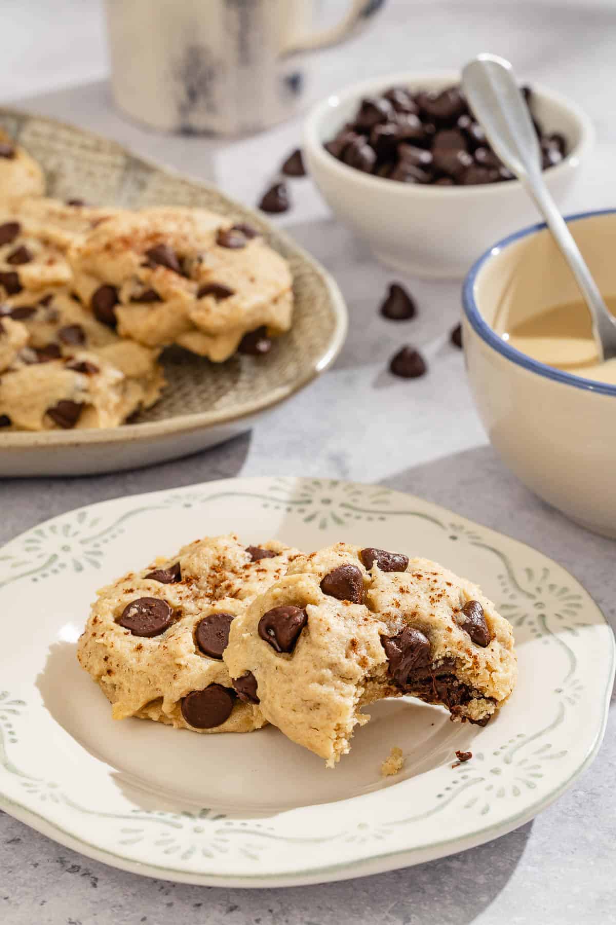 Two chocolate chip cookies on a plate, one with a bite taken out of it. Next to this is a platter with the rest of the cookies, bowls of tahini and chocolate chips and a mug of coffee.