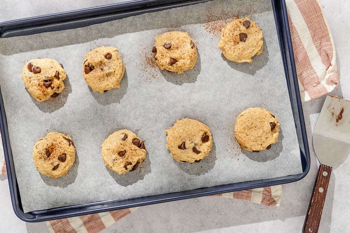 Eight tahini chocolate chip cookies on a parchment lined baking sheet resting on a kitchen towel next to a spatula.