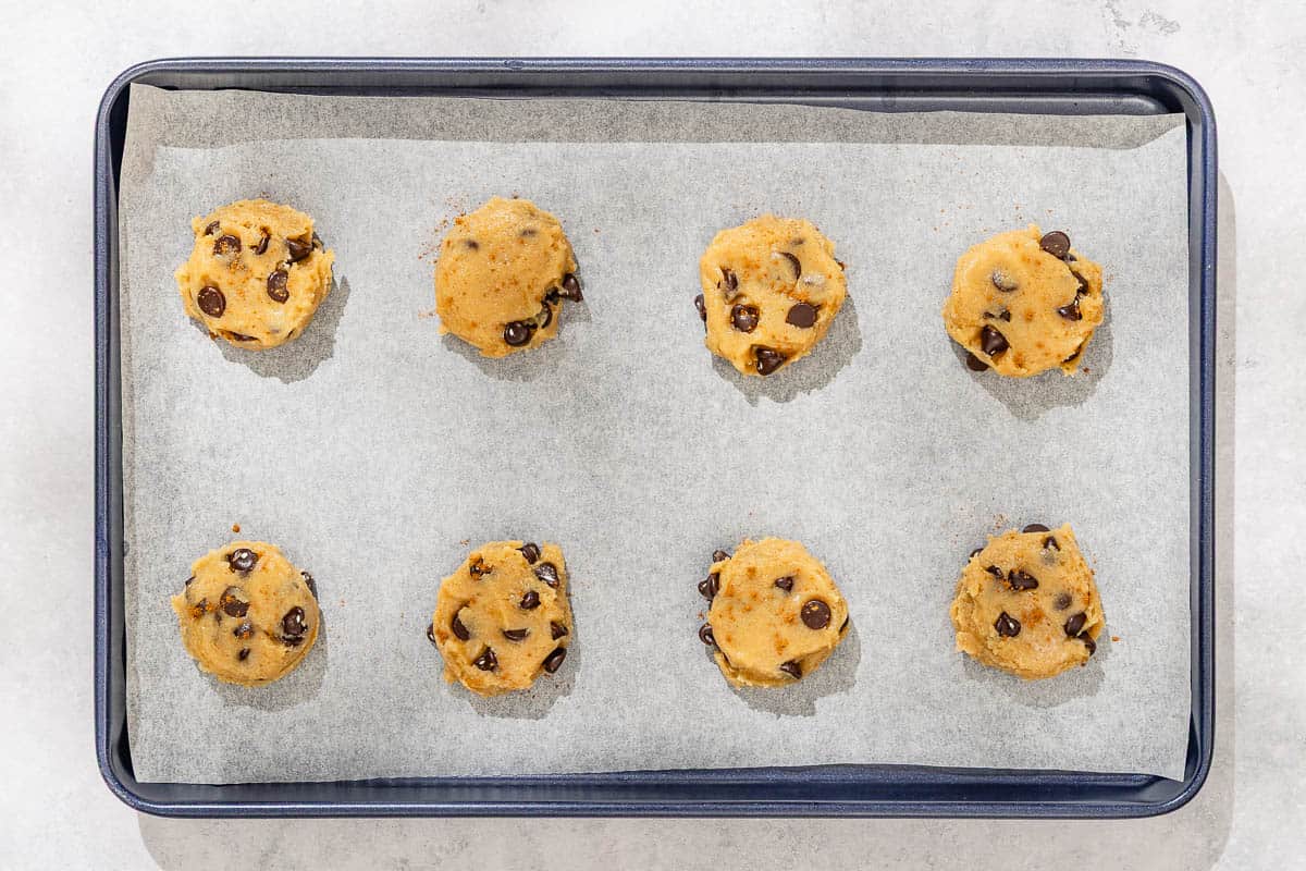 Eight slightly flattened unbaked tahini chocolate chip cookie dough balls spread evenly on a parchment lined sheet pan.