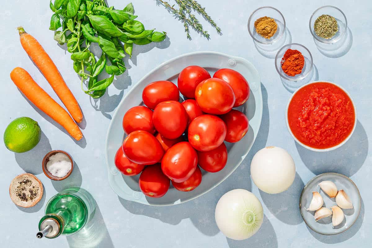 Ingredients for tomato basil soup including tomatoes, carrots, olive oil, salt, pepper, onions, garlic, canned crushed tomatoes, fresh basil, fresh thyme, oregano, paprika, cumin and lime.