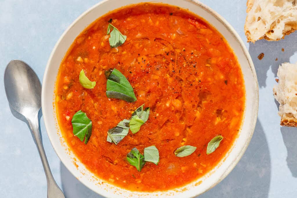 A bowl of tomato basil soup next to a spoon and pieces of crusty bread.