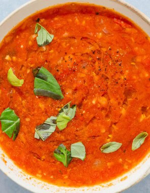 A bowl of tomato basil soup next to a spoon and pieces of crusty bread.