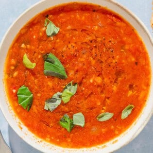 A bowl of tomato basil soup next to a spoon and pieces of crusty bread.