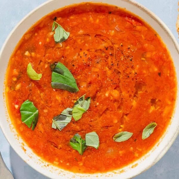 A bowl of tomato basil soup next to a spoon and pieces of crusty bread.