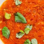 A close up of a bowl of tomato basil soup with pieces of crusty bread in the background.