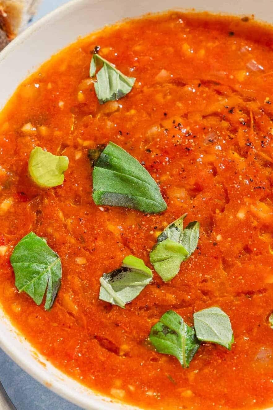 A close up of a bowl of tomato basil soup with pieces of crusty bread in the background.