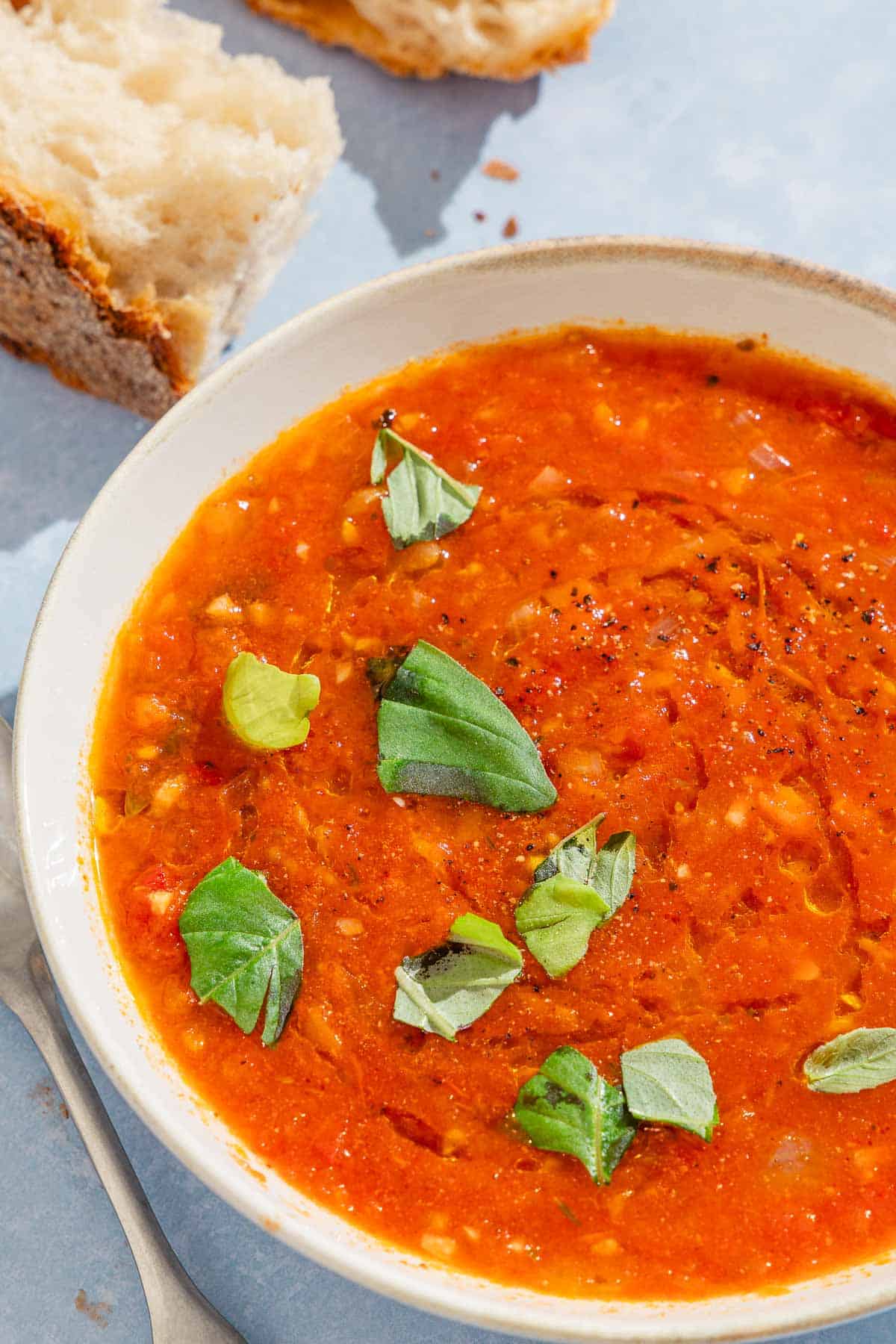 A close up of a bowl of tomato basil soup with pieces of crusty bread in the background.
