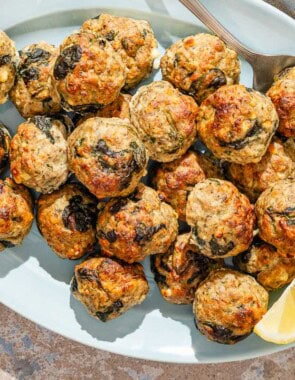 Several turkey meatballs on a platter with a serving spoon and lemon wedges. Next to this is a bowl of chopped parsley.