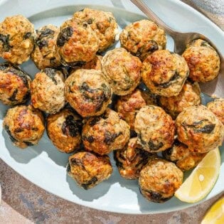 Several turkey meatballs on a platter with a serving spoon and lemon wedges. Next to this is a bowl of chopped parsley.