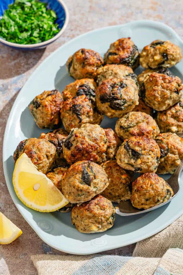 Several turkey meatballs on a platter with a serving spoon and lemon wedges. Next to this is a bowl of chopped parsley.
