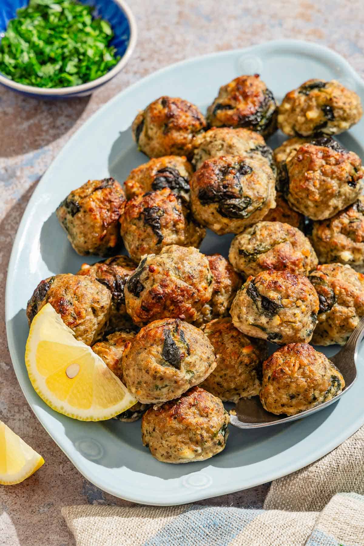 Several turkey meatballs on a platter with a serving spoon and lemon wedges. Next to this is a bowl of chopped parsley.