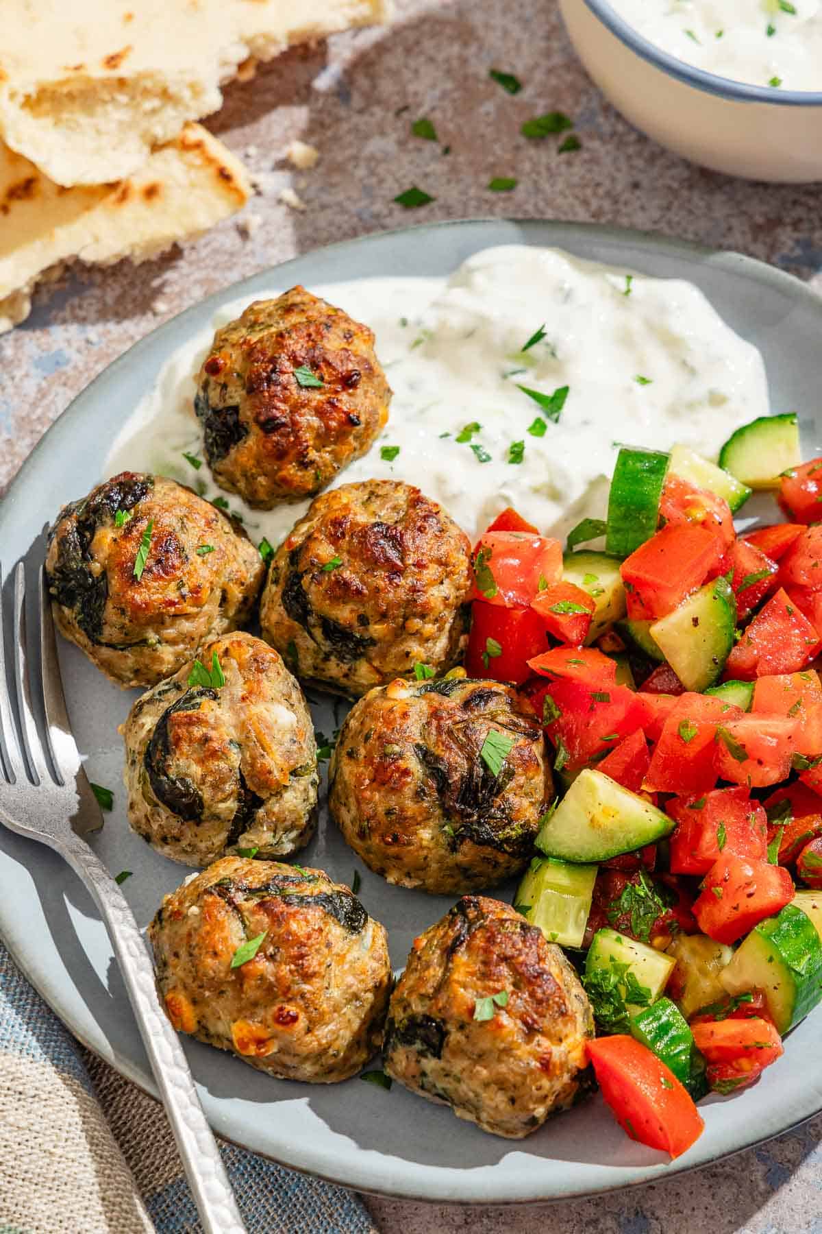 Several turkey meatballs on a plate with a fork, tzatziki sauce and a tomato cucumber salad. Next to this is more tzatziki sauce in a bowl, and pieces of pita bread.