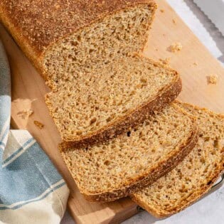 A partially sliced whole wheat bread loaf behind 3 slices of the bread on a cutting board. Next to this is a kitchen towel and a knife.