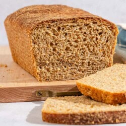 A partially sliced whole wheat bread loaf on a cutting board. Next to this is a knife, 2 slices of the bread and a kitchen towel.