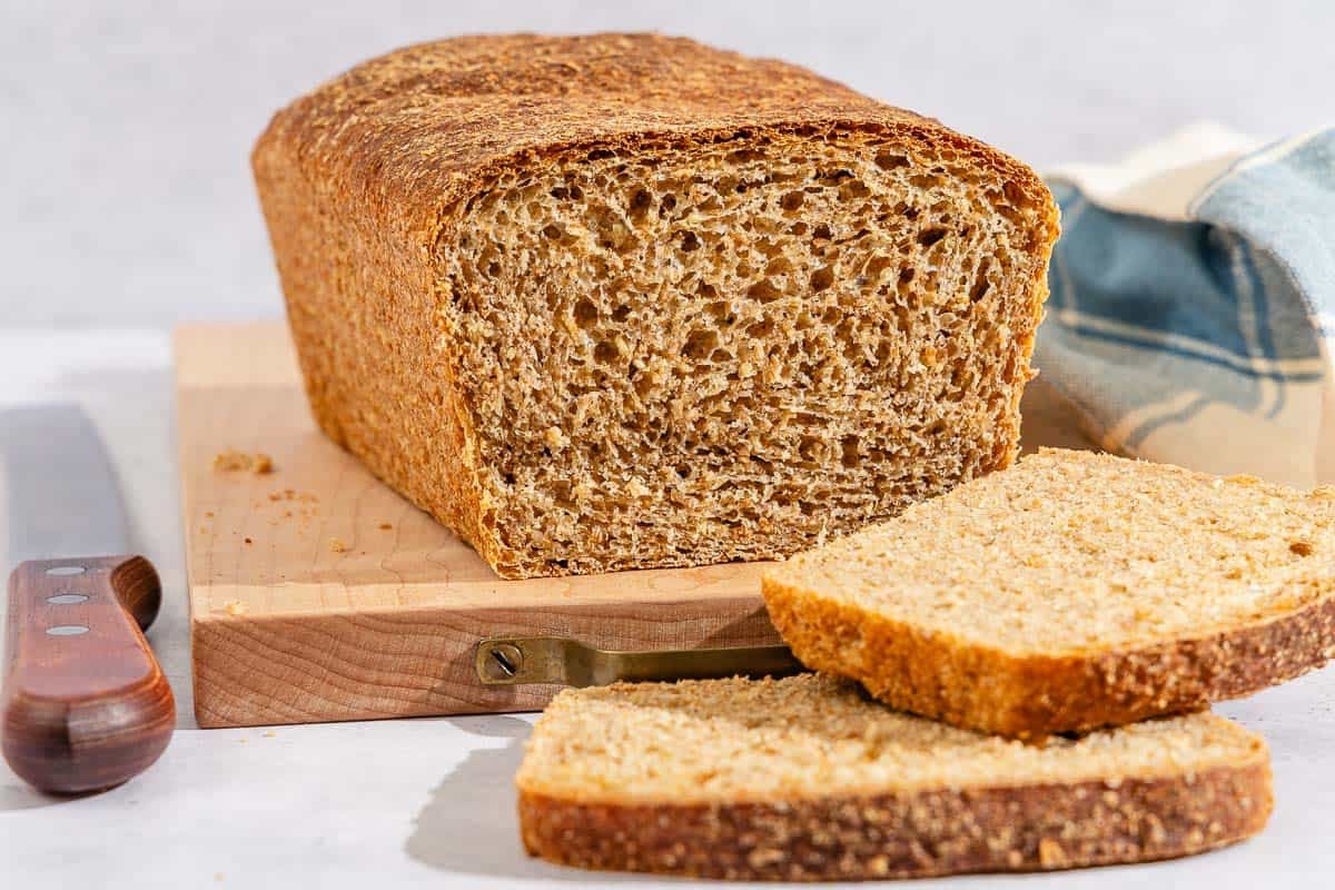 A partially sliced whole wheat bread loaf on a cutting board. Next to this is a knife, 2 slices of the bread and a kitchen towel.