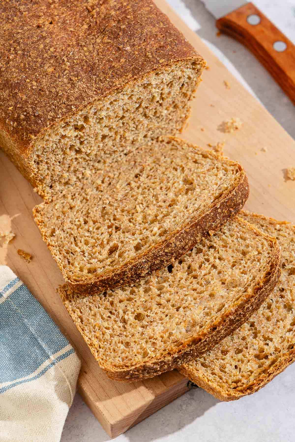 A partially sliced whole wheat bread loaf behind 3 slices of the bread on a cutting board. Next to this is a kitchen towel and a knife.