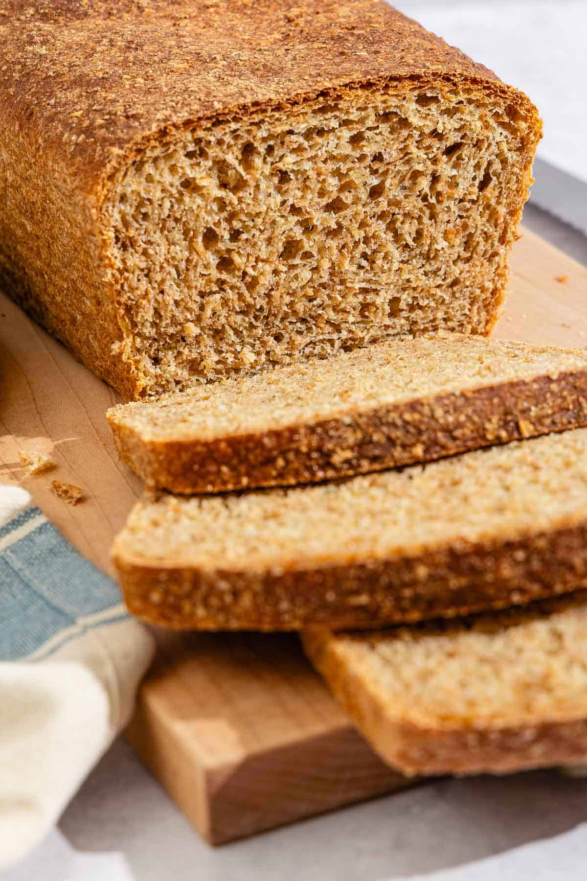 A partially sliced whole wheat bread loaf behind 3 slices of the bread on a cutting board.